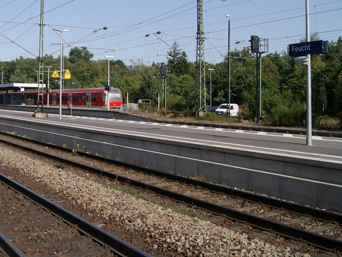 Bahnschienen und ein roter Zug, auf einem blauen Bahnhofsschild steht in weißer Schrift &quot;Feucht&quot;